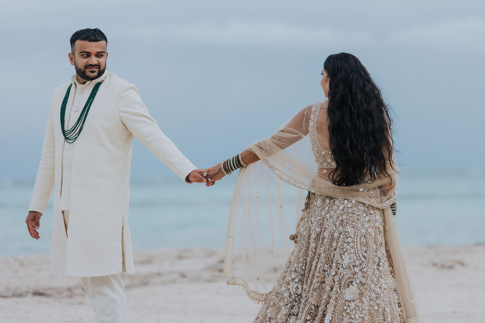 Bride and groom walking hand in hand along the beach in wedding attire