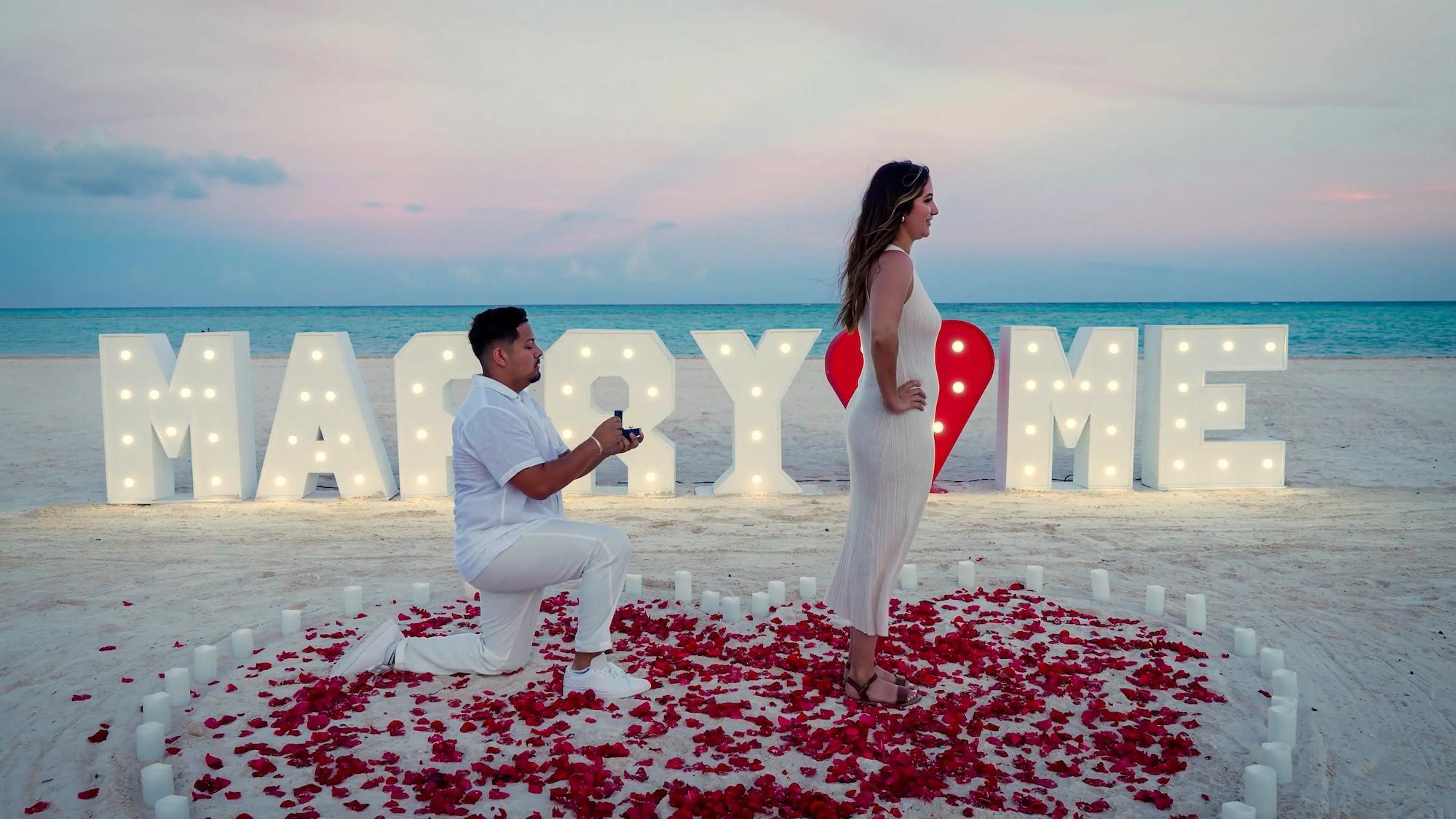 Beach proposal at dusk with illuminated MARRY ME letters and rose petals