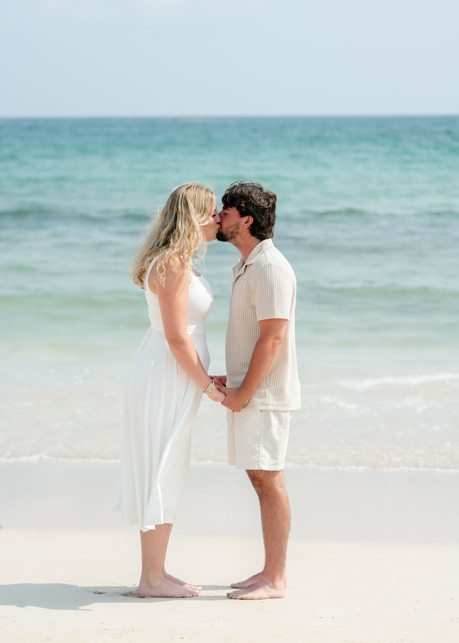 A newly engaged kiss barefoot at the shoreline
